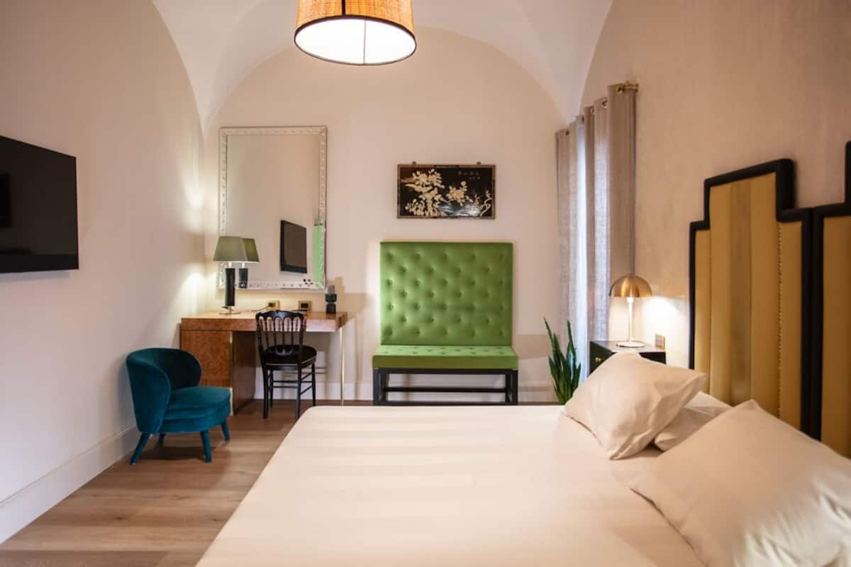Hotellihuone, jossa on suuri sänky, vihreäverhoiltu pääty, työpöytä tuolilla, televisio ja lamppu.