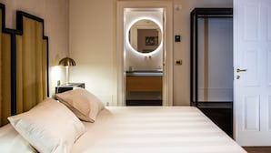 Habitación de hotel con cama, mesita de noche, lámpara, espejo con luz, puerta y vistas al baño.