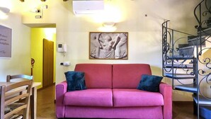 A living room with a red sofa, two dark cushions, a wooden dining table, and a staircase with a wrought iron railing.