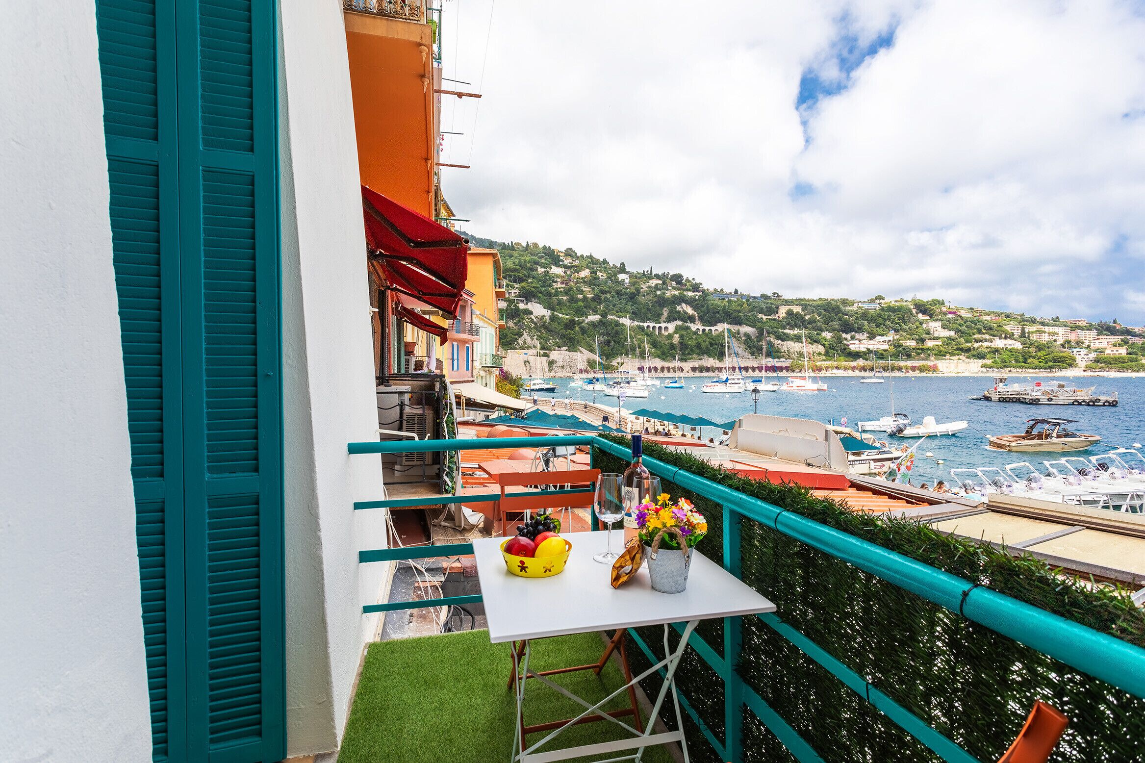 Un balcon avec une table blanche, un panier de fruits et un vase de fleurs. La vue comprend une marina avec des bateaux et une colline parsemée de bâtiments.