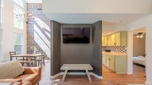 A modern living room with a large TV, a dining area, and a kitchen with green cabinets.