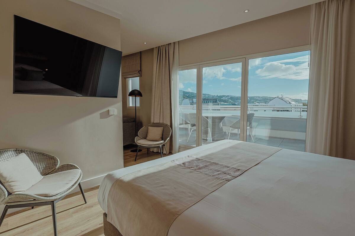 Une chambre d’hôtel avec un grand lit, un téléviseur fixé au mur, une chaise et une vue sur la ville grâce à de grandes fenêtres.