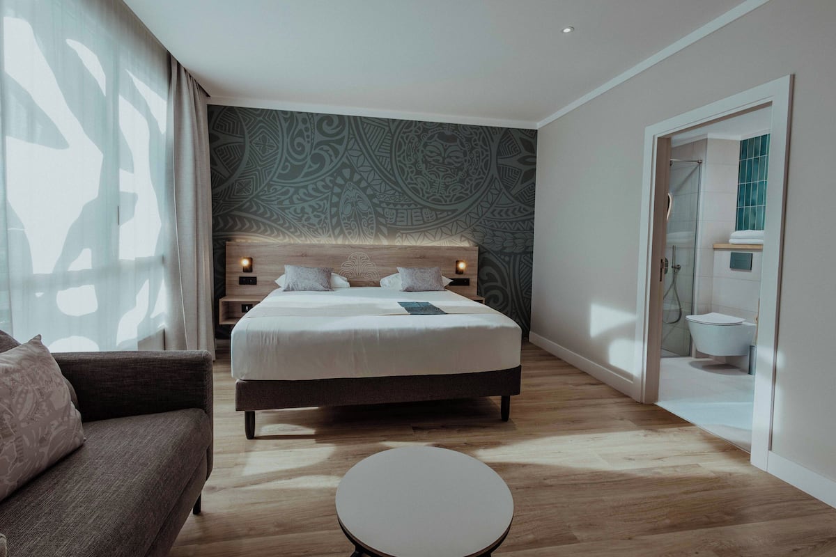 Une chambre d’hôtel moderne avec un grand lit, une petite table ronde, un canapé et un mur décoré de motifs.