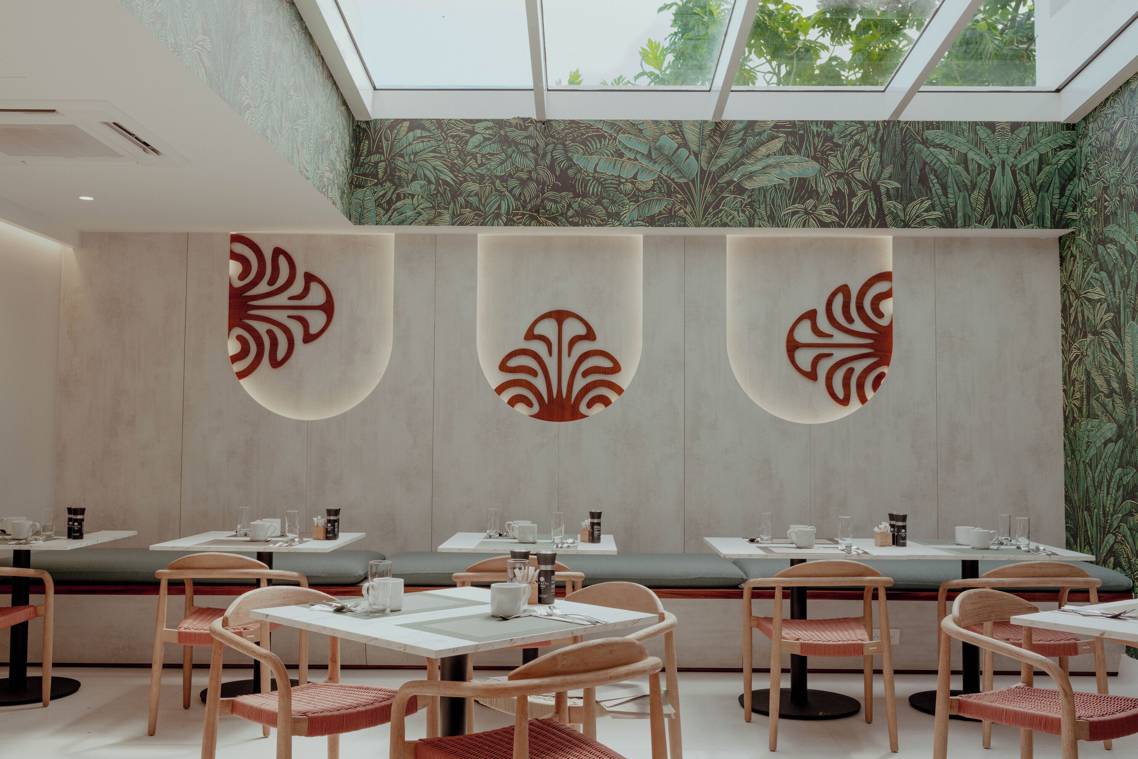 A modern restaurant with high ceilings, a skylight, and a green leafy wallpaper. Tables are set with white tablecloths and wooden chairs.