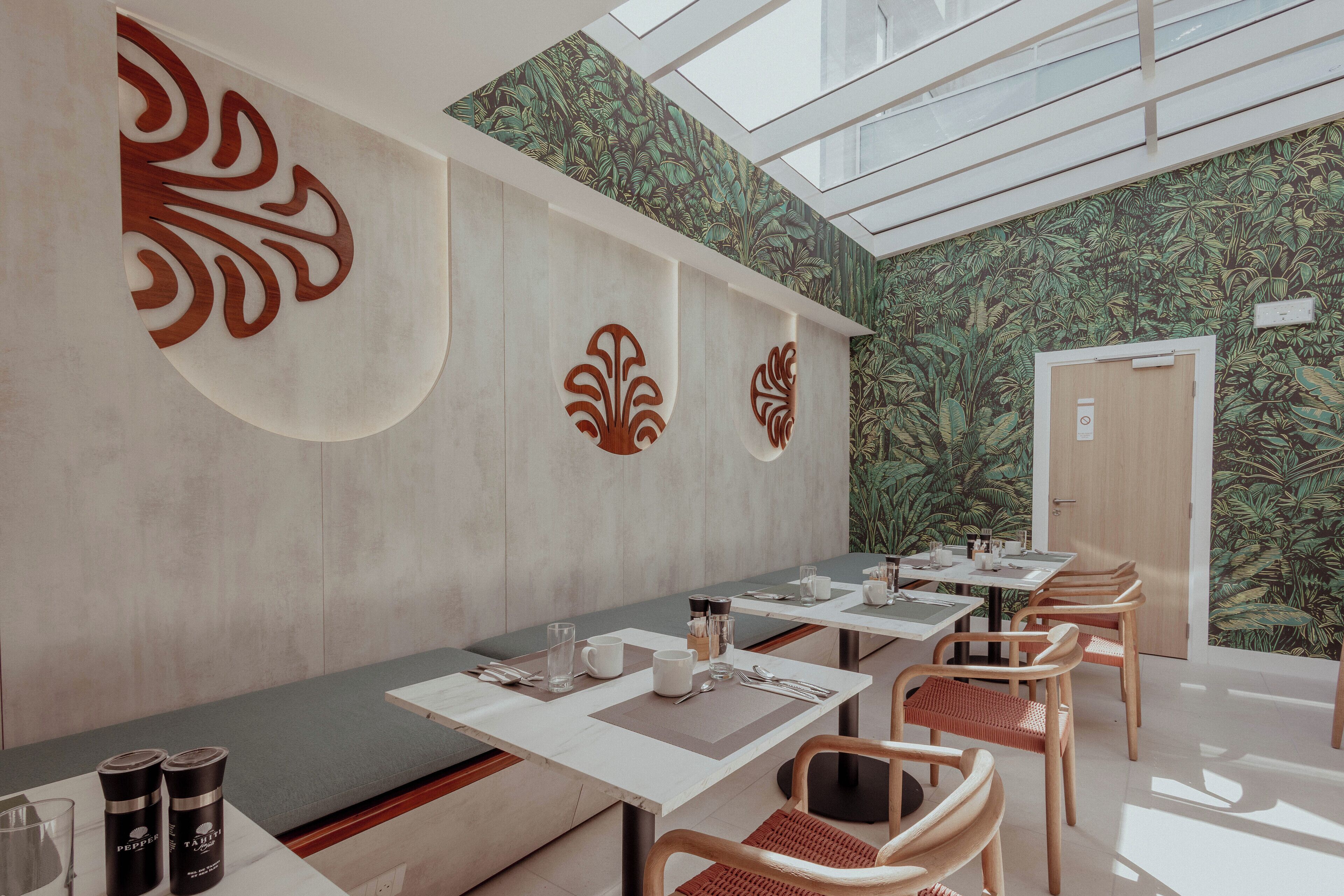 Ein modernes Restaurant mit hoher Decke, Oberlicht und tropischem Wanddesign. Die Tische sind mit weißen Tischdecken gedeckt, und es stehen Holzstühle bereit.