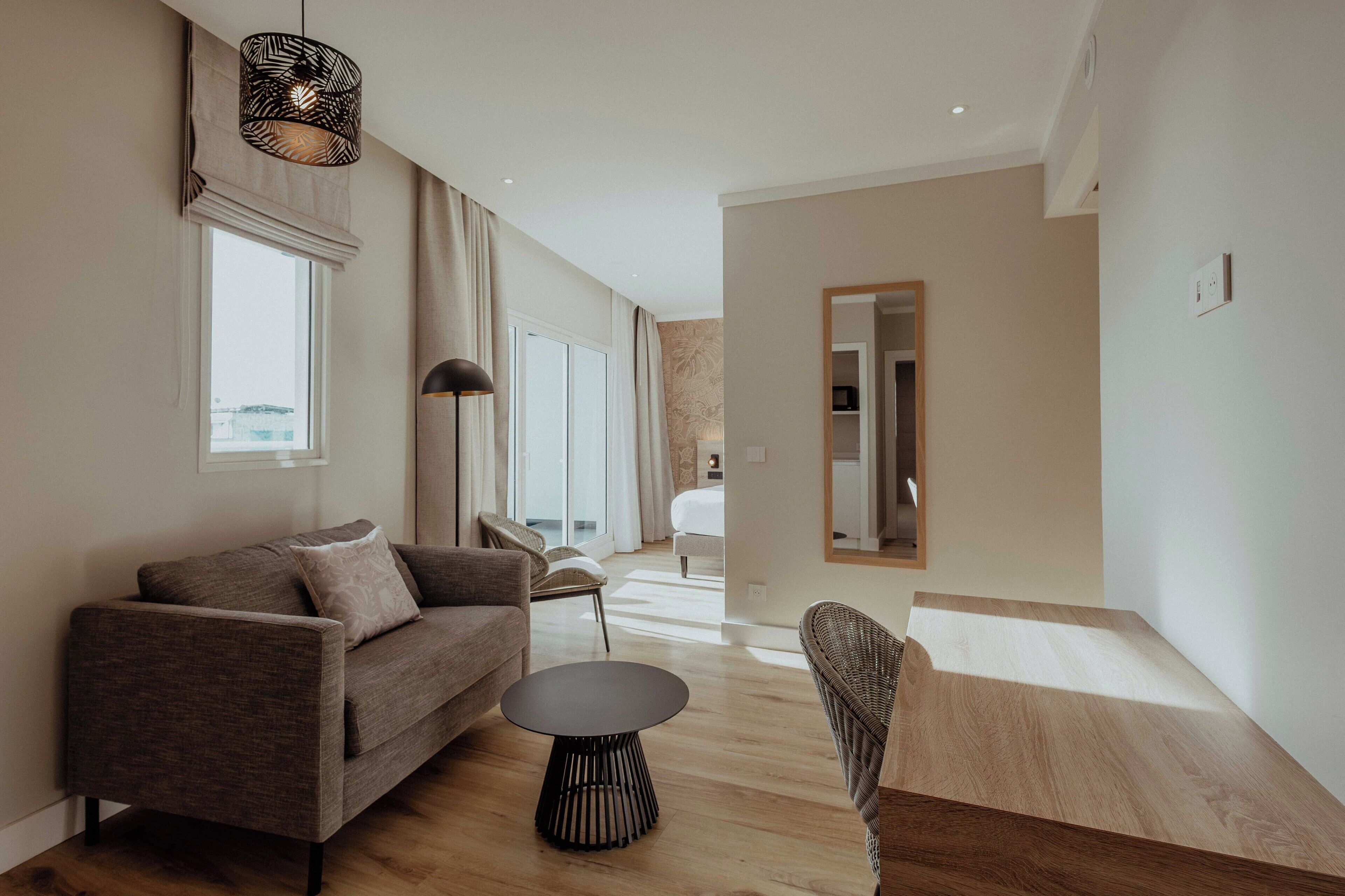 Een moderne woonkamer met een bank, een rond salontafeltje en een rieten stoel. Er is een groot raam met gordijnen, een plafondlamp en een spiegel aan de muur.