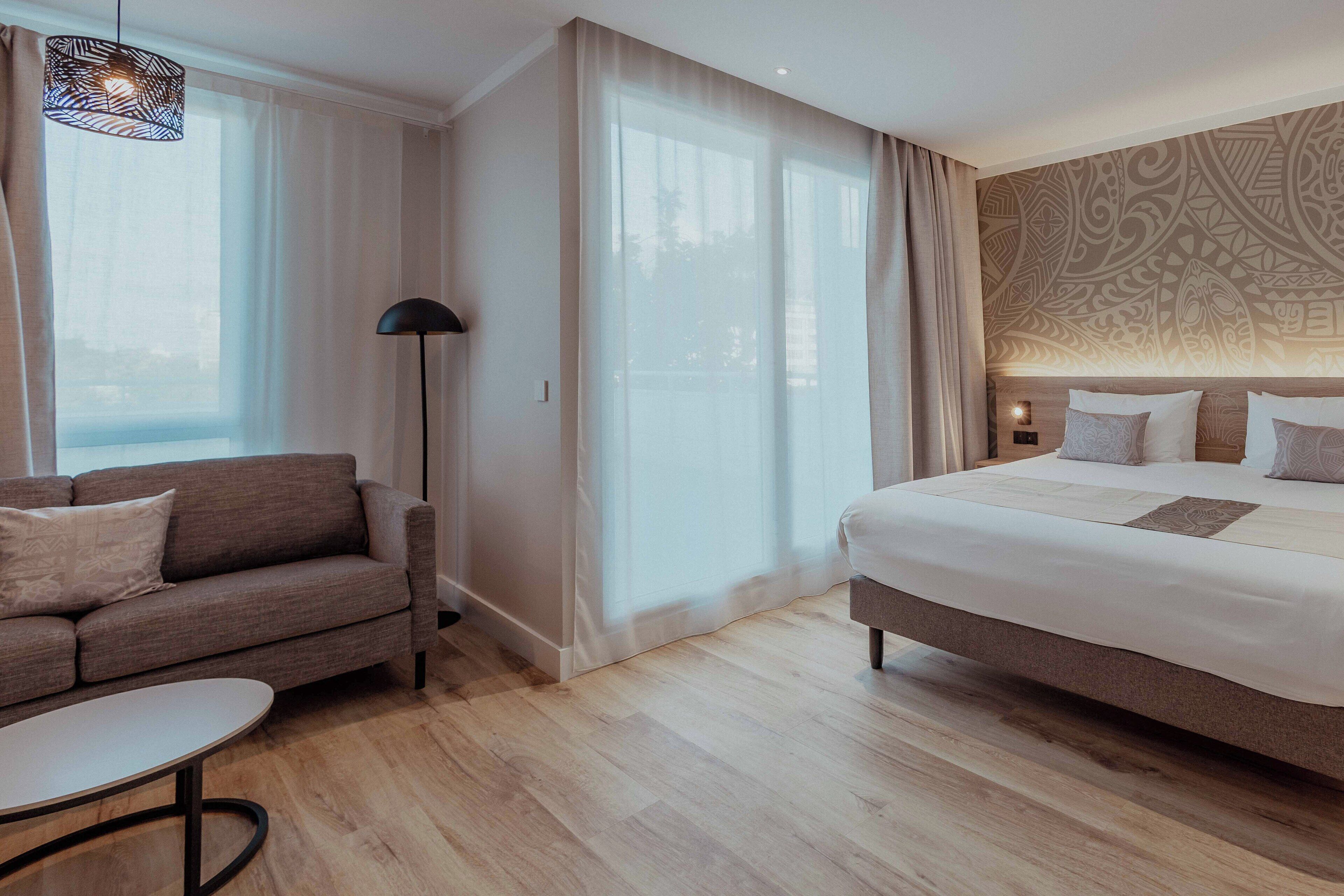 A modern hotel room with a large bed, a sofa, a small table, and a floor lamp.