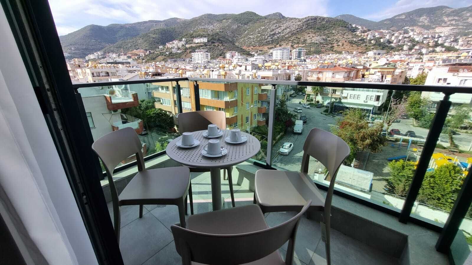 Un balcón con mesa y sillas, con vistas a un paisaje urbano que incluye edificios y montañas.