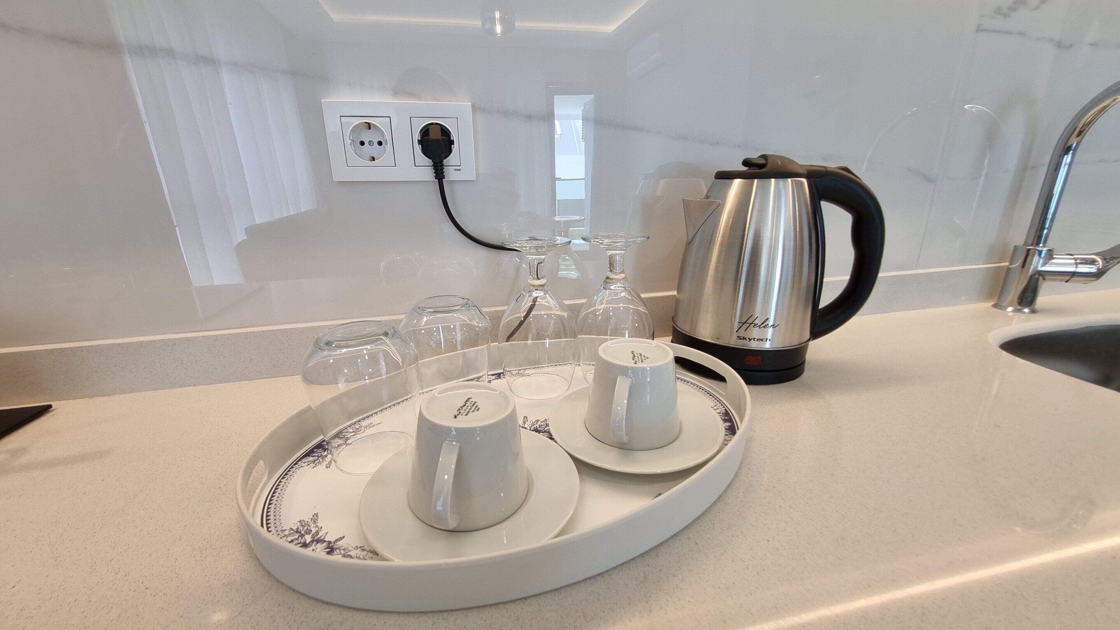 Un comptoir de cuisine avec un plateau contenant des tasses et des soucoupes blanches, une bouilloire et une prise électrique.