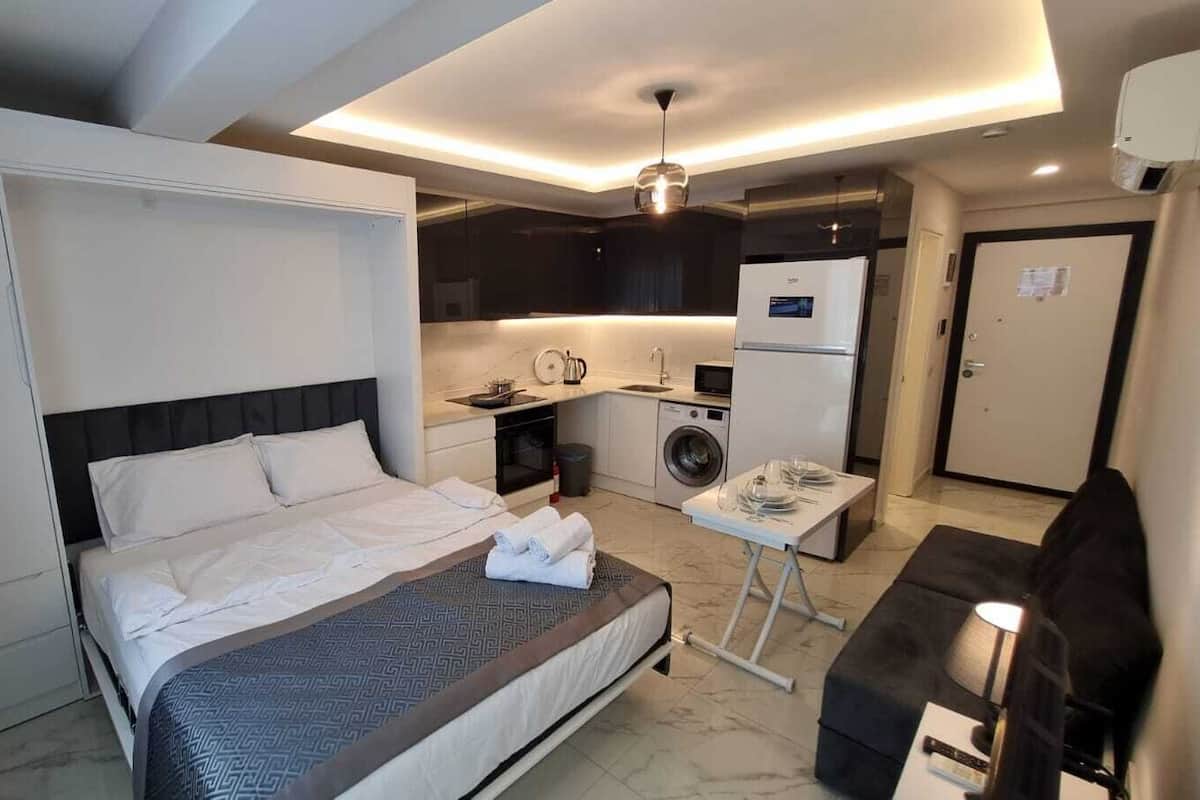 A modern hotel room with a large bed, a small kitchenette, a washing machine, and a compact dining area.
