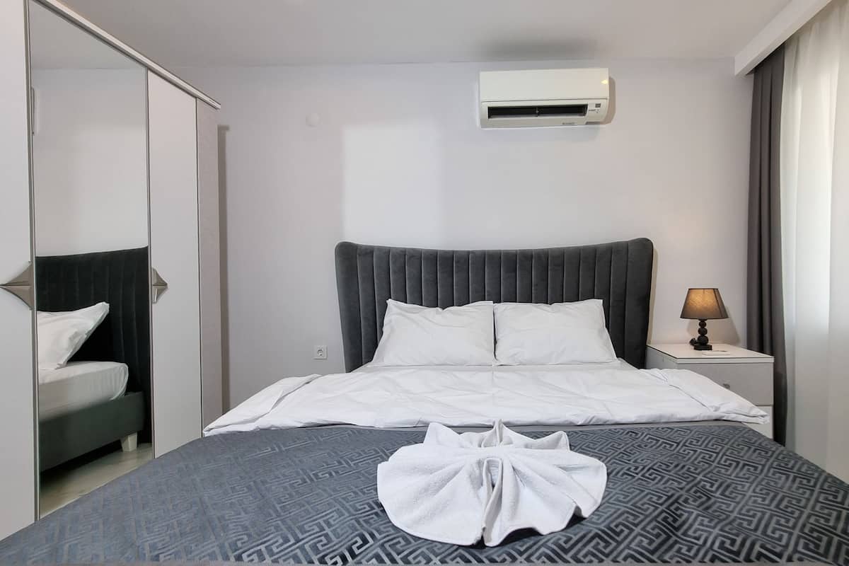 A hotel room with a bed, a bedside table with a lamp, a wall-mounted air conditioner, and a window with curtains.