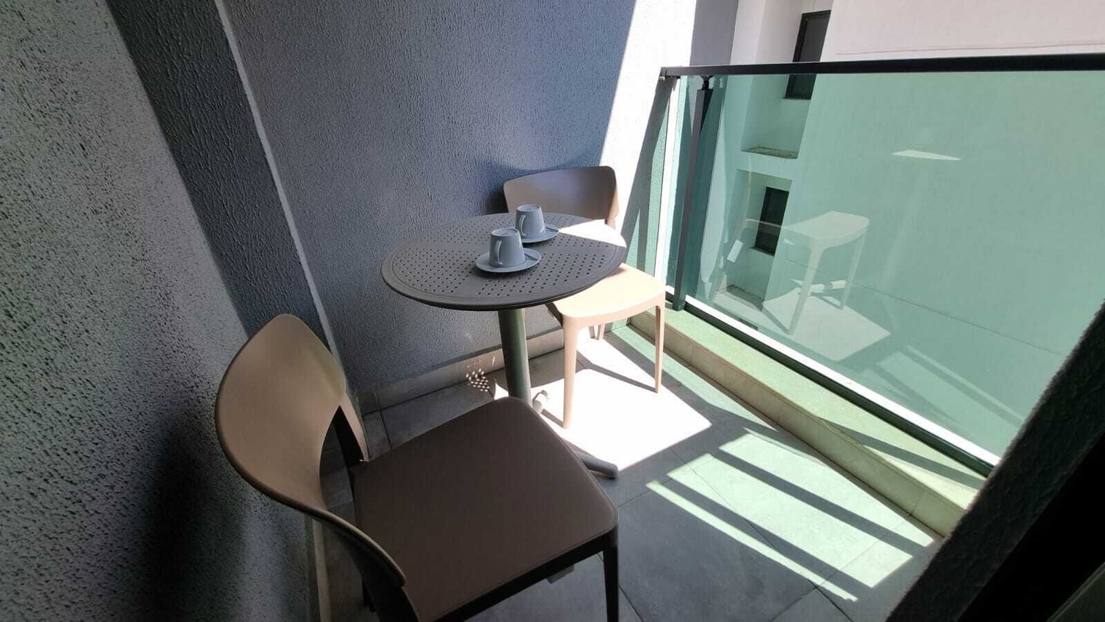 A balcony with a small table and two chairs, a glass railing, and a view of a building.