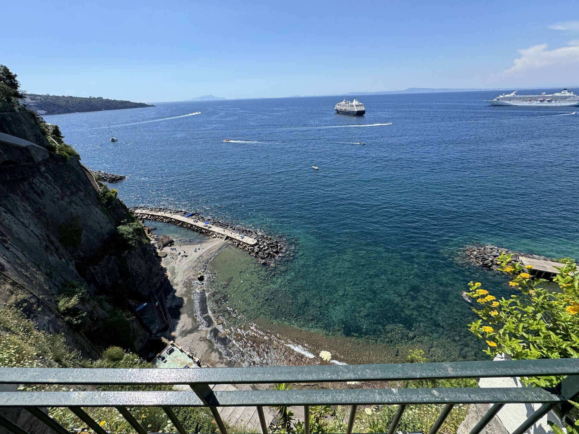A coastal view with a clear blue sea, a rocky shoreline, and a pier extending into the water.