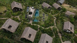 Vista aérea de un complejo turístico con cabañas de techo de paja, una piscina central y abundante vegetación.