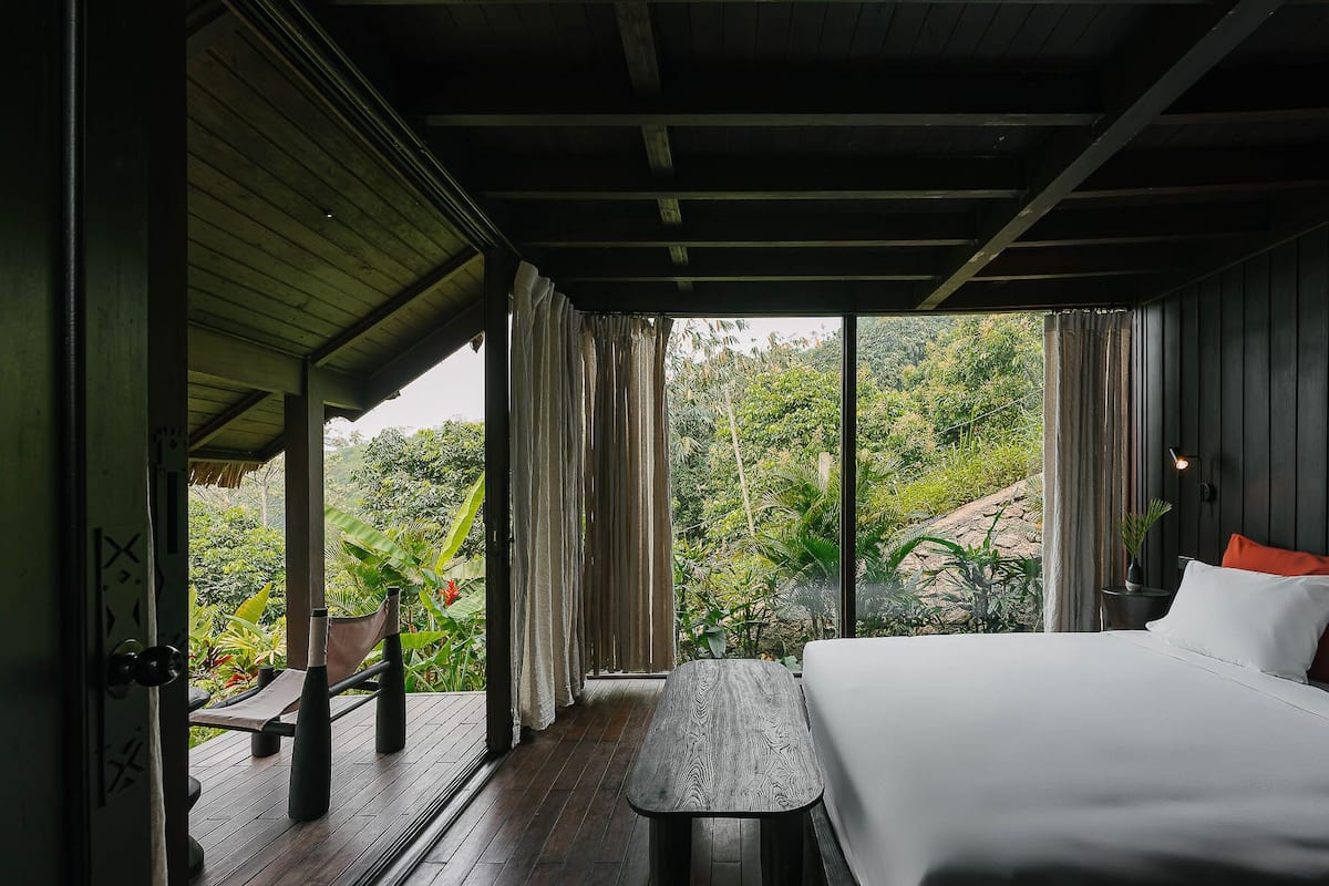 Une pièce avec un lit, une petite table et un banc. On peut voir de la verdure par la fenêtre.