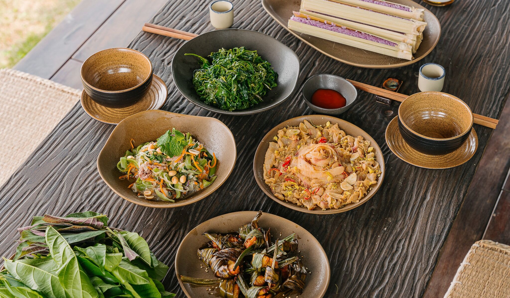 A table with various dishes, including a bowl of greens, a bowl of rice with shrimp, and a plate of wrapped items.