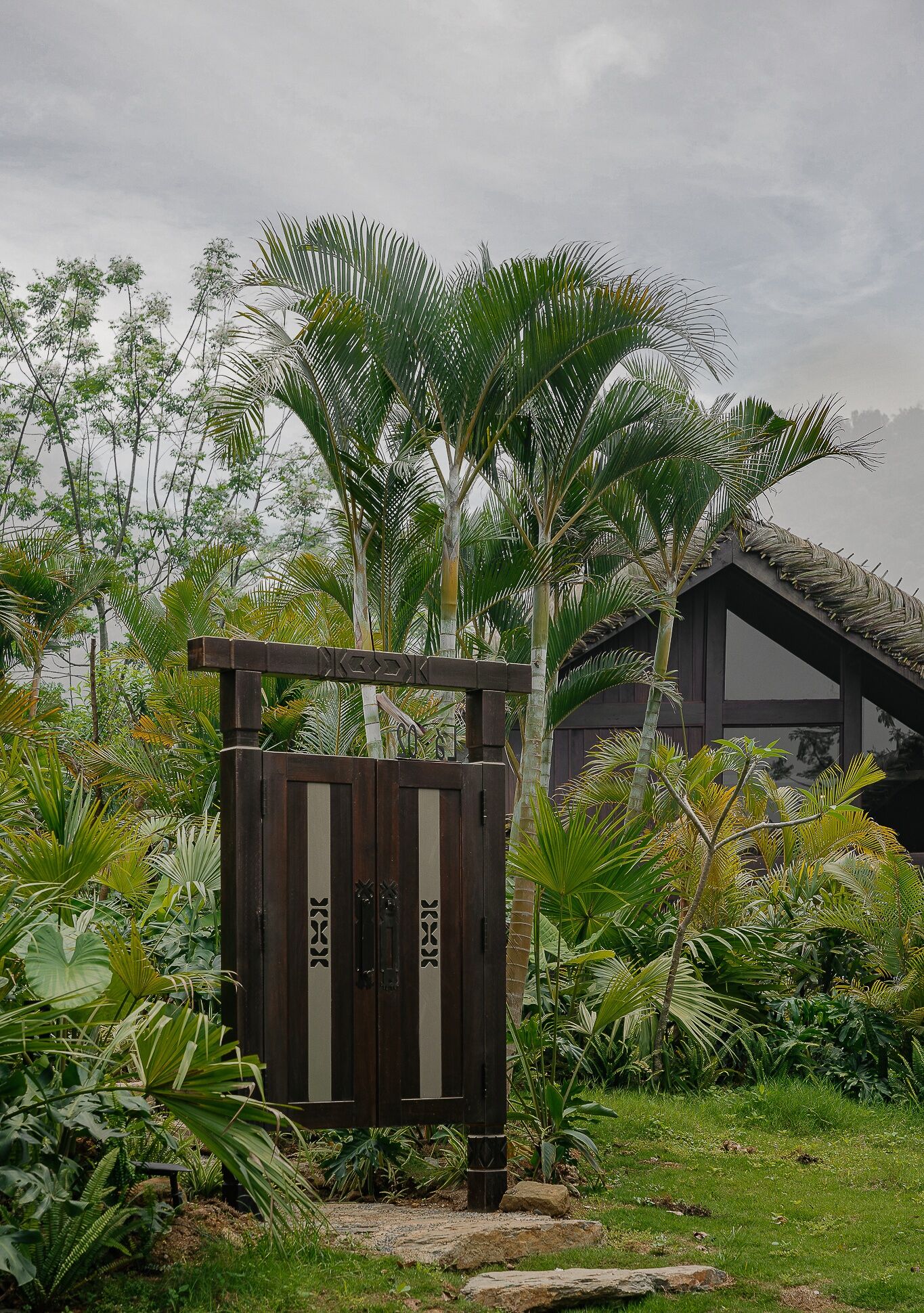 A wooden gate with vertical stripes, surrounded by lush tropical plants and a building with a thatched roof in the background.