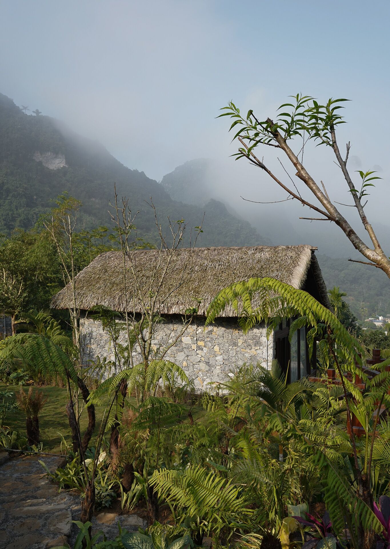 A rustic stone house with a thatched roof, surrounded by lush greenery and mountains in the background.