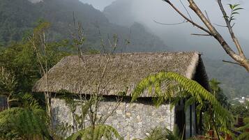 A rustic stone house with a thatched roof, surrounded by lush greenery and mountains in the background.
