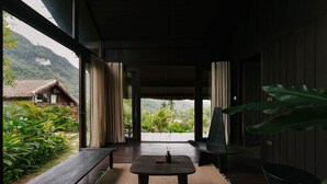 Una habitación con piso de madera, una mesa con un florero y vista a una montaña y un edificio con techo de paja.