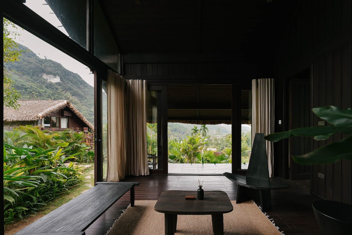 Une pièce avec un plancher en bois, une table ornée d’un vase, et une vue sur une montagne et un bâtiment à toit de chaume.