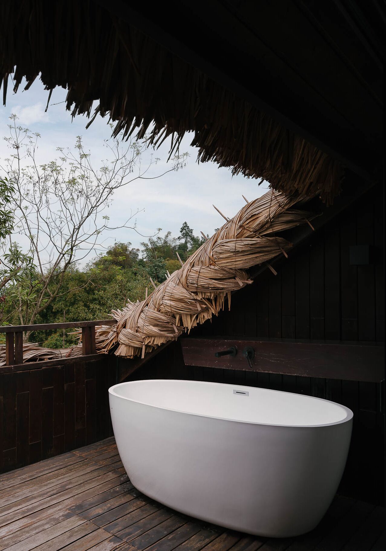 A large, white bathtub under a thatched roof structure, surrounded by wooden decking and a wooden fence.