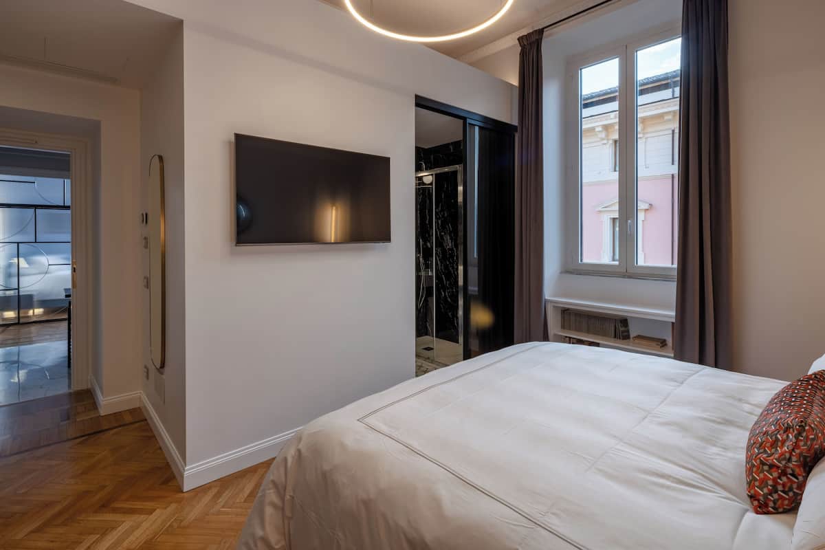 A modern bedroom with a large bed, a flat-screen TV mounted on the wall, and a view of a building through a window.