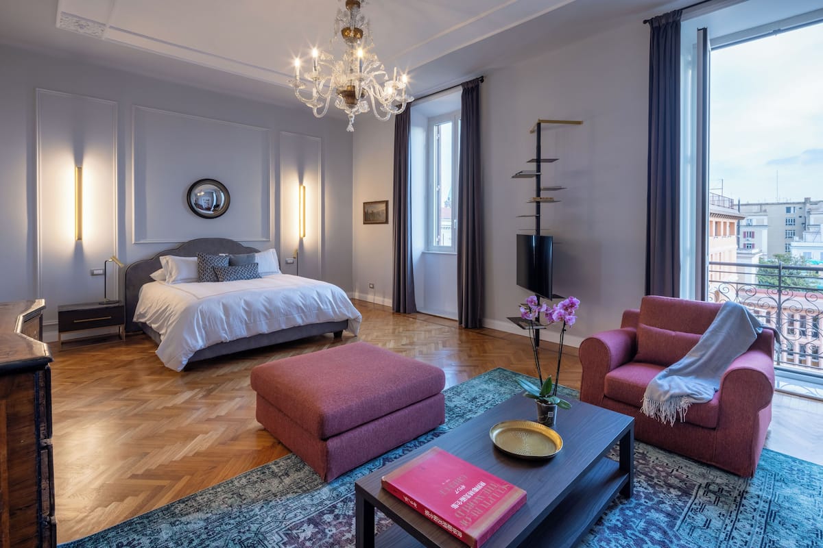 A modern bedroom with a large bed, a sitting area with a sofa and armchair, a coffee table with a book, and a view of the city through large windows.