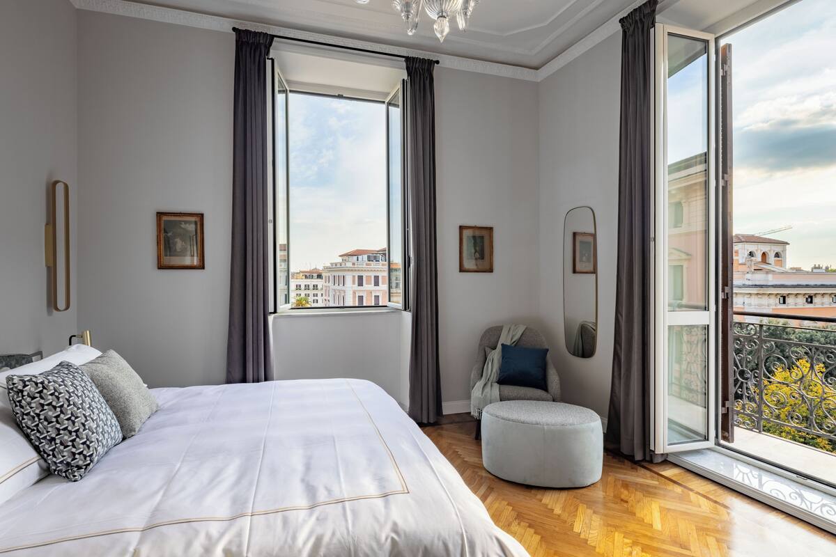 A modern bedroom with a large bed, a round ottoman, and a view of the city through a large window.