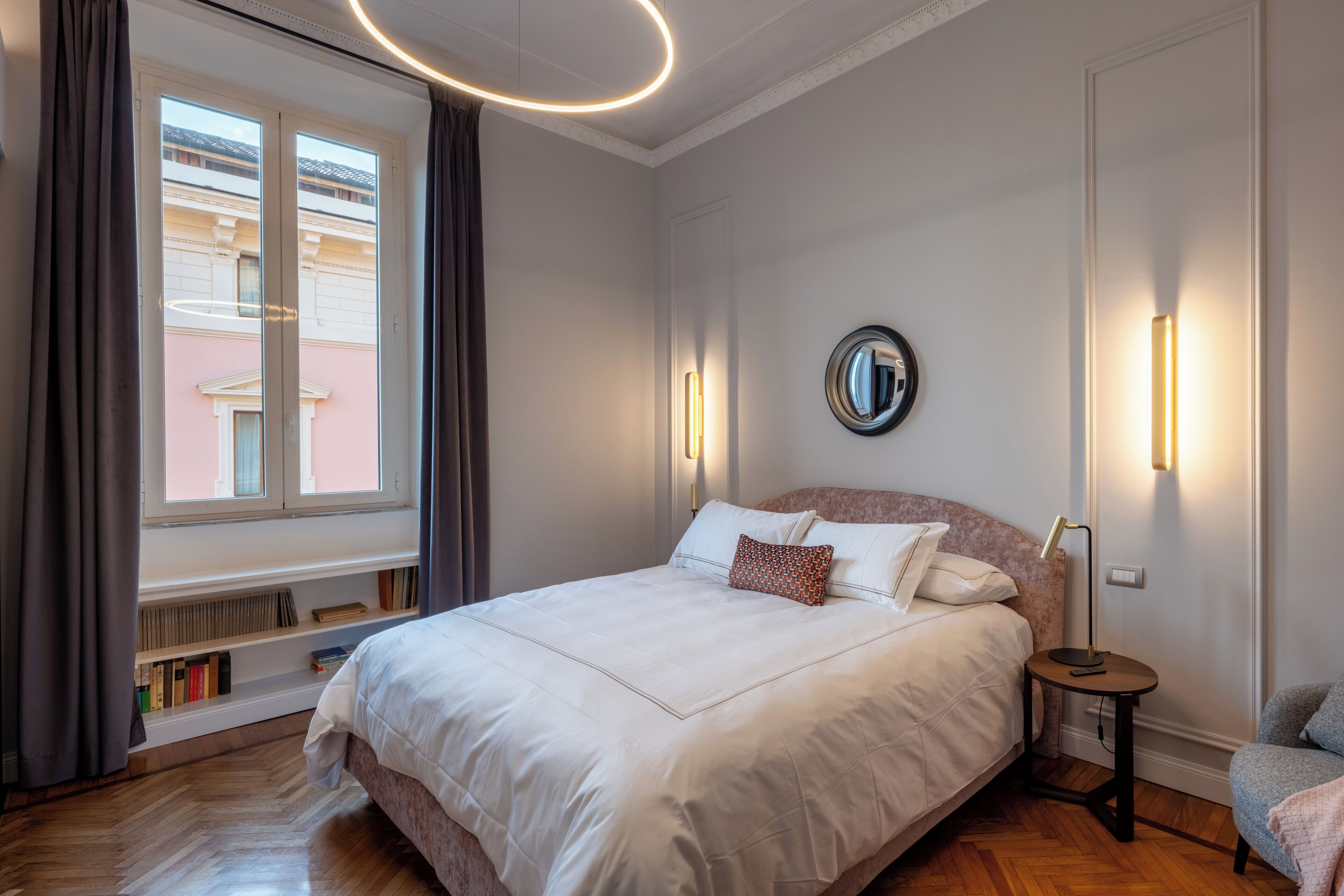 A bedroom with a large bed, bedside lamps, a round mirror, and a window with curtains.