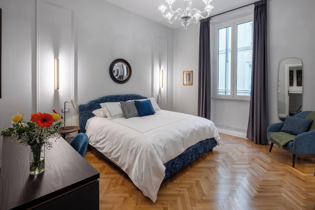 A modern bedroom with a large bed, a mirror, a desk with a vase of flowers, and a view of the city through a window.