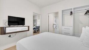 Une chambre d’hôtel moderne dotée d’un grand lit, d’un téléviseur à écran plat fixé au mur, d’un dressing et d’une salle de bain visible à travers une porte ouverte.