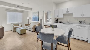 Una sala de estar moderna y bien iluminada con una mesa de comedor redonda, sillas tapizadas en gris, un sofá color beige y un amplio ventanal con persianas.