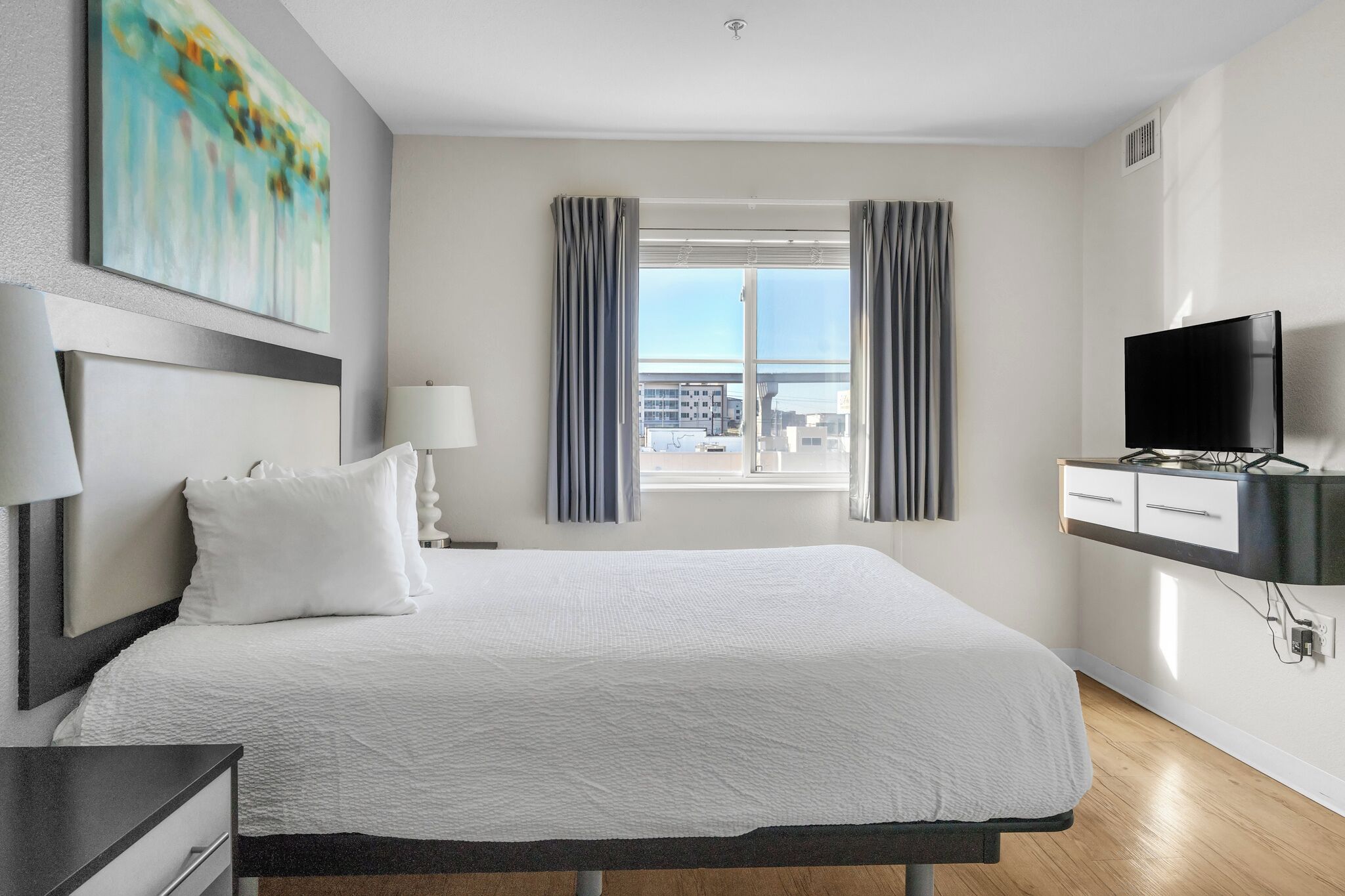 A modern hotel room with a large bed, a flat-screen TV, a window with curtains, and a city view.