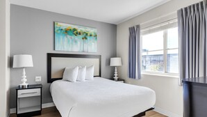 Ett modernt hotellrum med en stor säng, två sängbord med lampor, en tavla på väggen och ett fönster med gardiner.