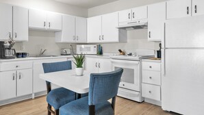 A modern kitchen with white cabinets, a white dining table with blue chairs, a microwave, and a refrigerator.