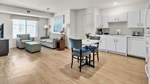 A modern living room with a sofa, armchairs, a dining table with chairs, a kitchen area with white cabinets, and a large window with blinds.