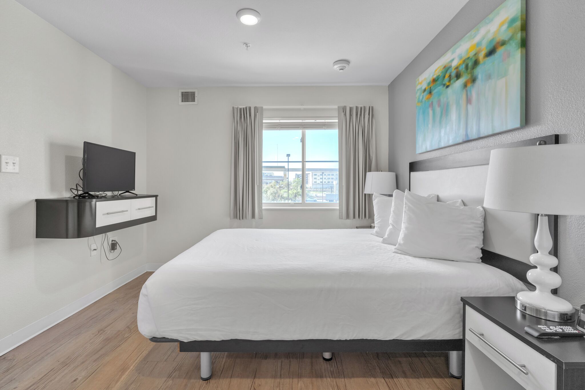 A modern hotel room with a large bed, a TV mounted on the wall, a bedside table with a lamp, and a window with curtains.