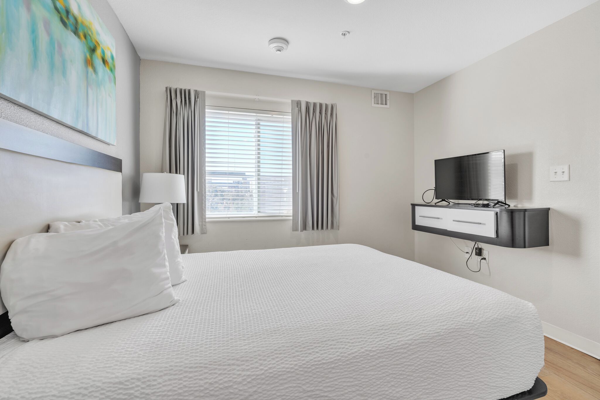 Een hotelkamer met een groot bed, een aan de muur bevestigde televisie, een raam met gordijnen en een schilderij.