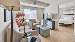 A modern hotel room with a sofa, armchair, ottoman, a flat-screen TV, a bed, and a potted plant. - Simplicity At the Rim 1216 I Comfy 2 Bed 2 Bath (San Antonio)