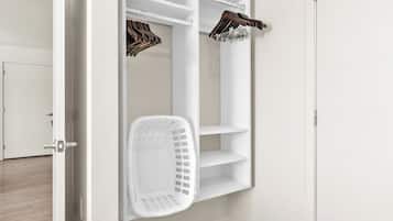 A white closet with shelves and a hanging rack, a white laundry basket, and a door.