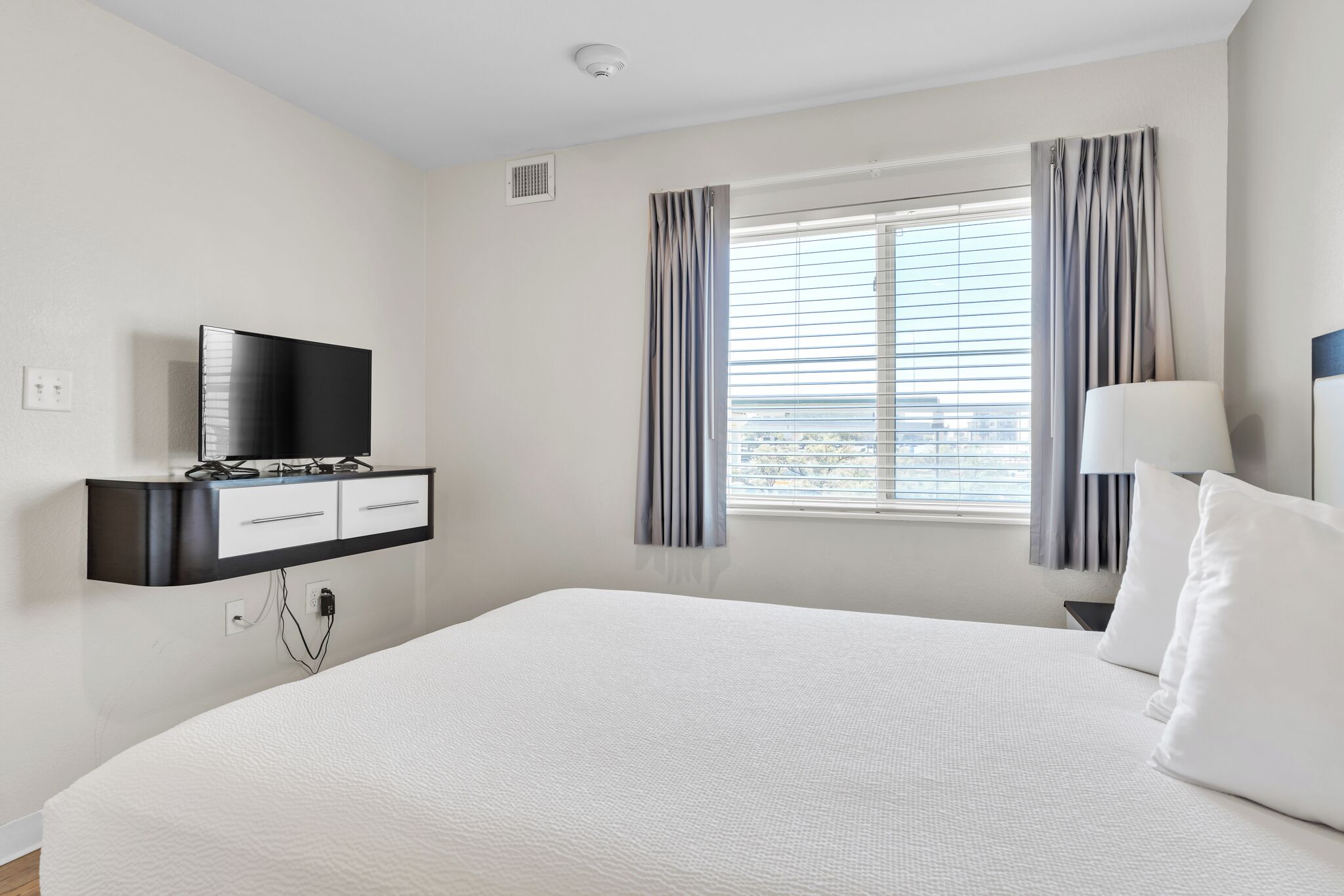 Een hotelkamer met een groot bed, een televisie bevestigd aan een zwart kastje, een raam met gordijnen en een bedlamp.