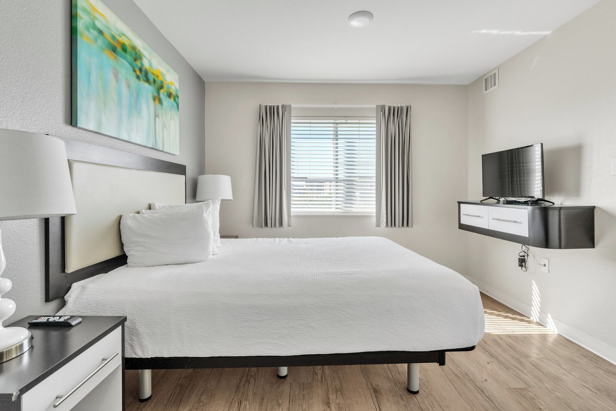 Una habitación de hotel moderna con una cama grande, un televisor de pantalla plana, un escritorio con una lámpara y un cuadro en la pared.