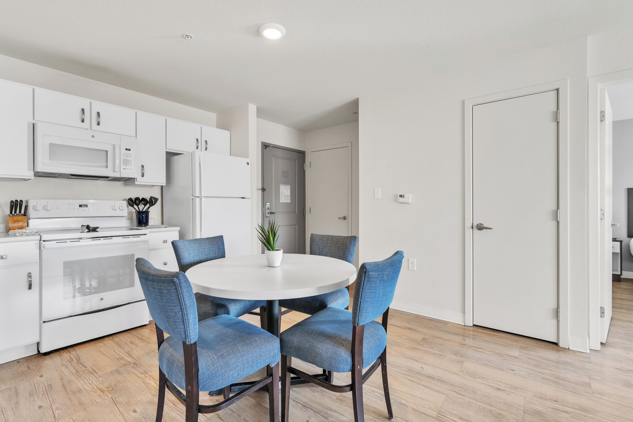 Una cocina moderna con electrodomésticos blancos, una mesa de comedor con sillas azules y una puerta que da a otra habitación.