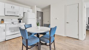 A modern kitchen with white appliances, a dining table with blue chairs, and a door leading to another room.
