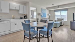 A modern kitchen with white cabinets, a dining area with blue chairs, and a large window with blinds.