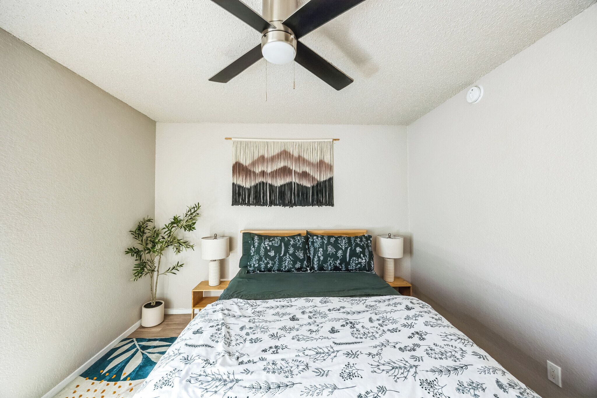 Ein Schlafzimmer mit einem Bett, einem Deckenventilator, einer Wandteppich, Nachttischlampen und einem Blumentopf.