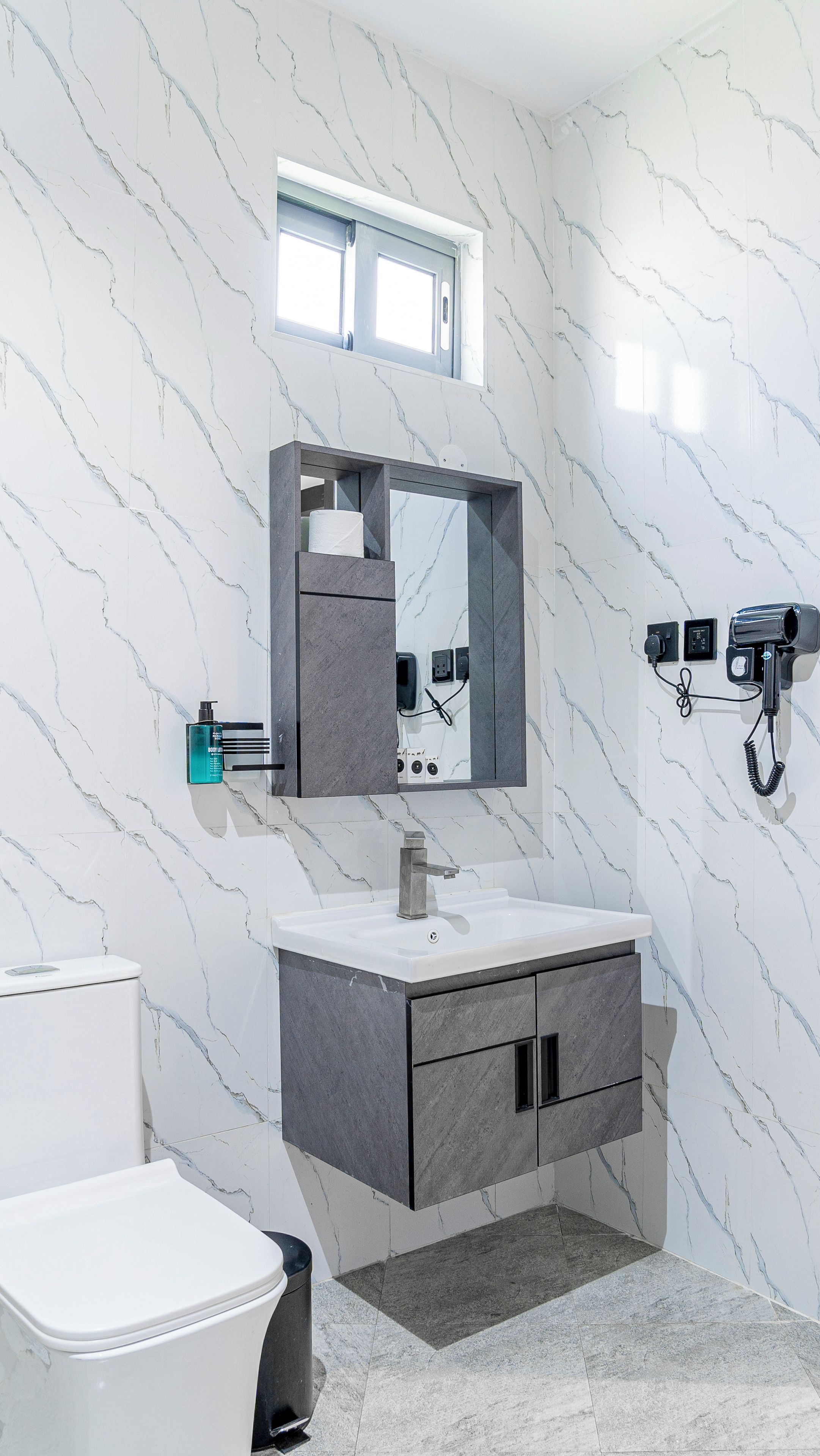A bathroom with a marble wall, a sink with a mirror above it, a hairdryer on the wall, and a toilet.