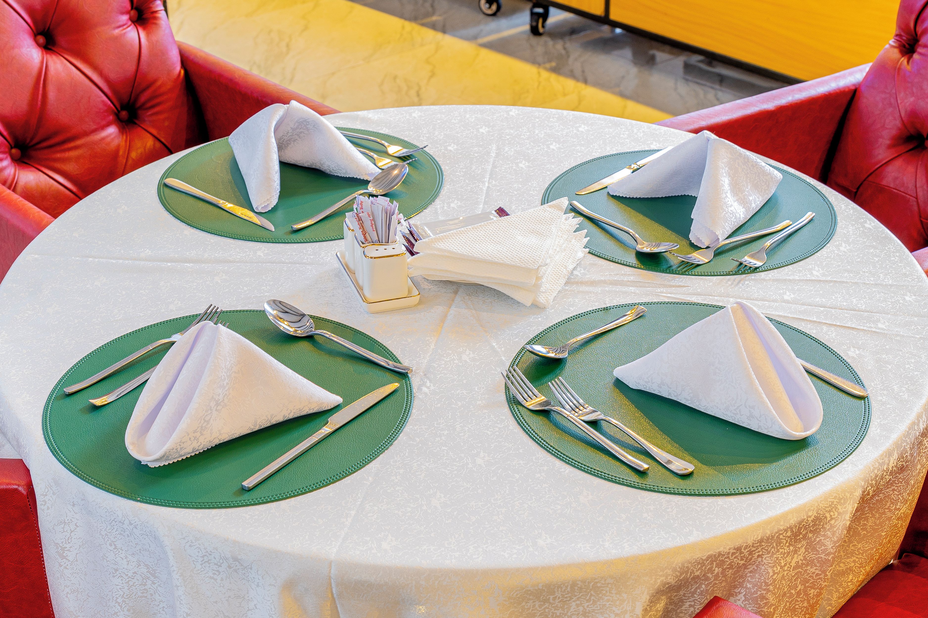 Mesa redonda para comer con individuales verdes y servilletas blancas dobladas en forma de conos, cubiertos y un pequeño jarrón blanco con flores.