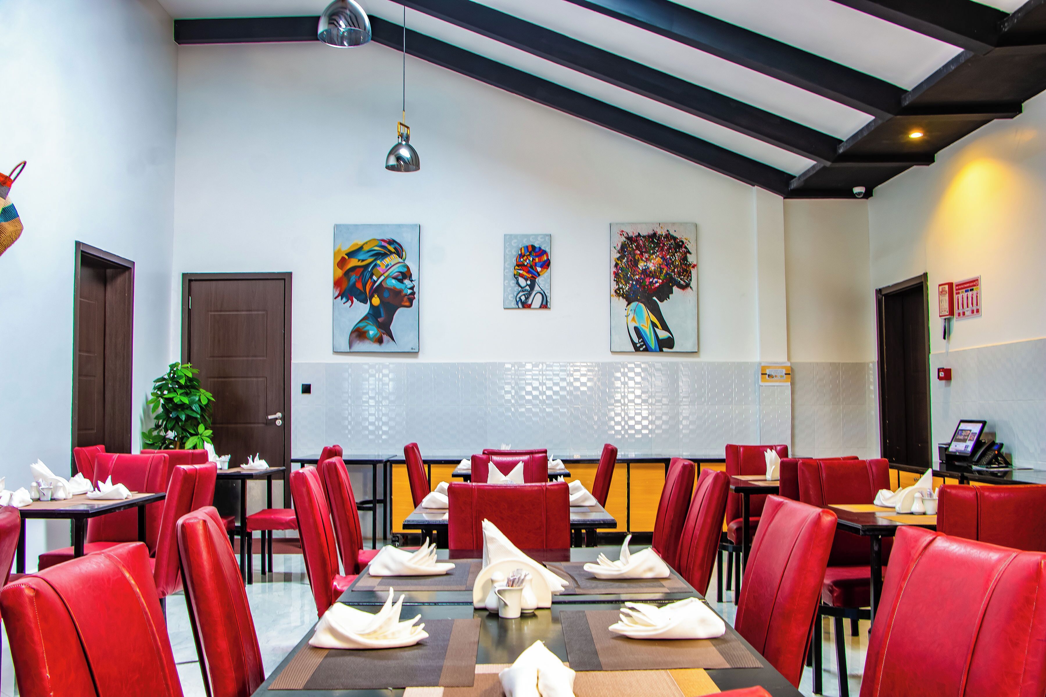 Un comedor con sillas tapizadas en rojo, mesas dispuestas con servilletas y platos blancos, y tres cuadros en la pared.